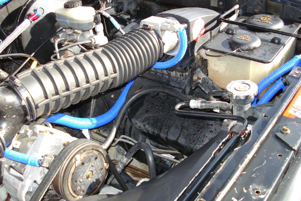 Engine bay showing large air intake tube, brake fluid reservoir, and blue corrugated hoses. Watermarks for Supermotors.net are visible