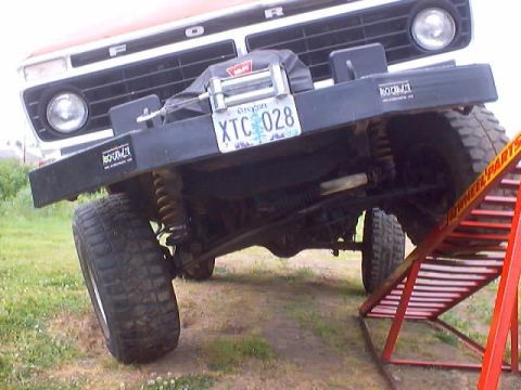 1976 Ford F-100 - Low front view of a lifted 1970s Ford pickup on grass with one front wheel flexed up onto a red ramp, exposing the solid front axle, coil s…