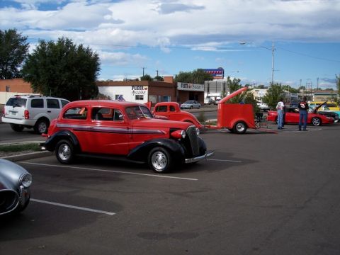 1972 Ford Ranchero - A red vintage sedan with black fenders and chrome trim is parked at a car show, with other vehicles and buildings in the background.
