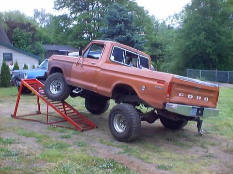 1976 Ford F-100 - Orange Ford F-100 pickup truck is elevated on a red ramp, with a blue truck visible behind it.