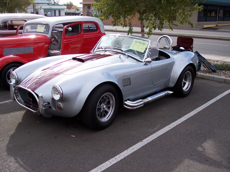 Silver convertible sports car with red stripes and side exhaust pipes parked next to a red hot rod