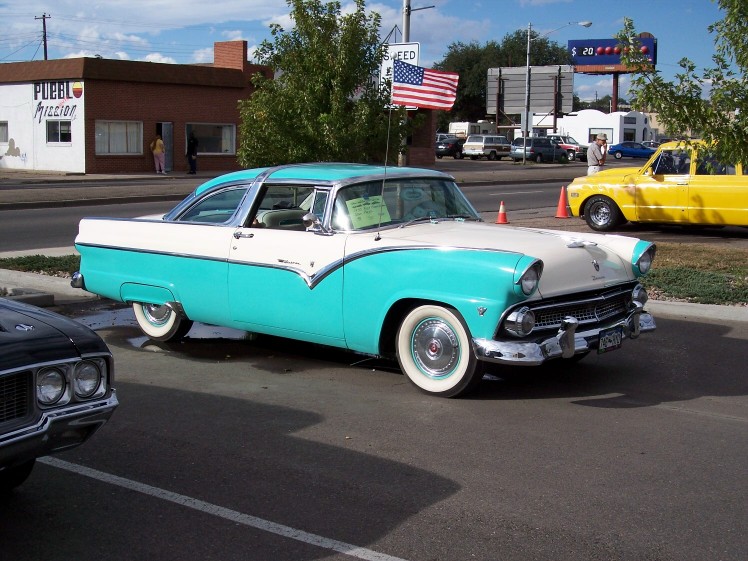 Turquoise and white 1955 Ford Fairlane Crown Victoria parked curbside, with a yellow pickup truck in the background