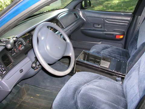 1999 Ford Crown Victoria - The interior of a police car shows a console unit between the front seats, with the steering wheel and dashboard visible.