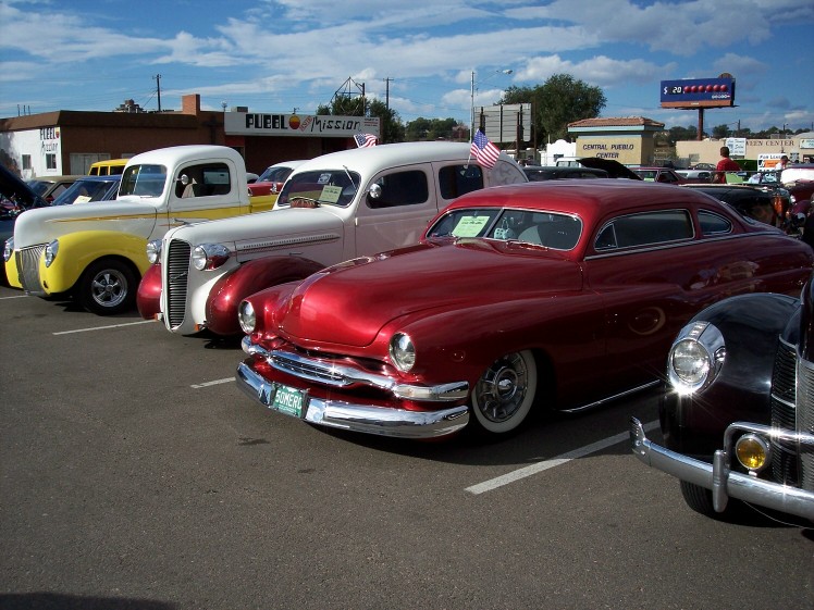 Red 1950s coupe with wide chrome grille and bumper, parked at a car show