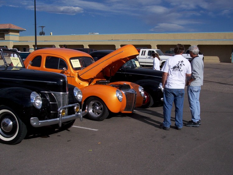 Two guys check out an orange hot rod with its hood up, parked next to a black classic car