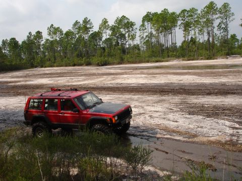 1989 Ford Bronco - Red Jeep Cherokee XJ navigating a challenging off-road terrain with muddy patches and sandy soil.