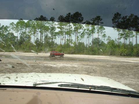 1989 Ford Bronco - A red SUV navigates a muddy terrain, with another vehicle's hood visible in the foreground.