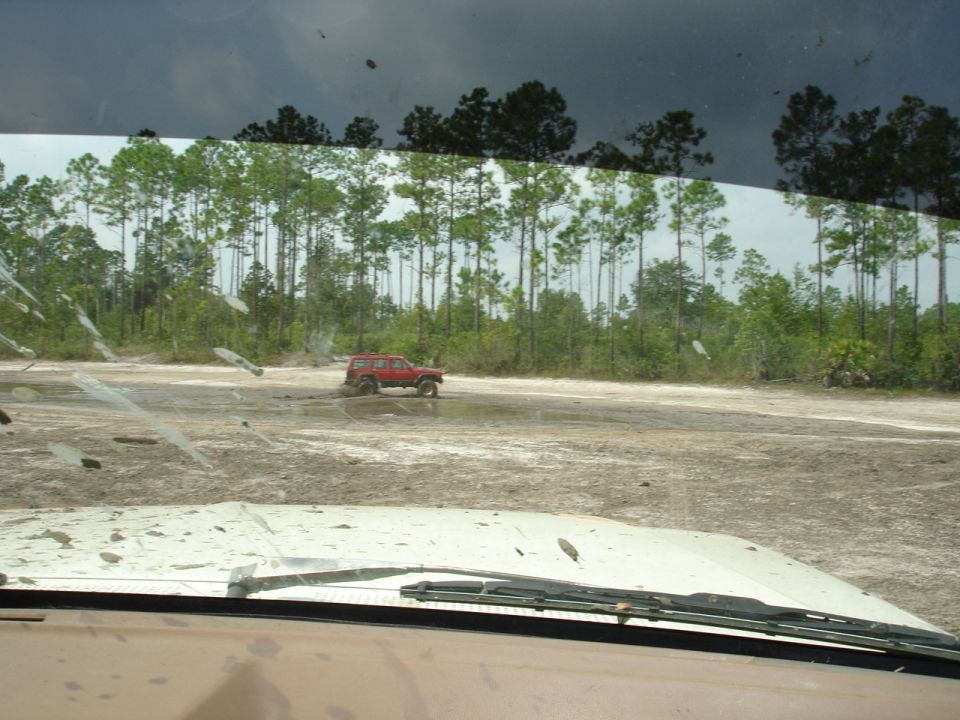 Red SUV with mud splattered on its side, driving through a muddy field with trees in the background