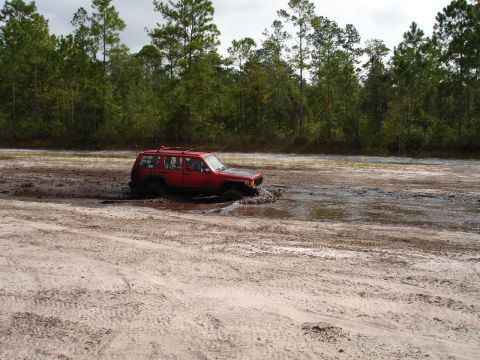1989 Ford Bronco - A red Jeep Cherokee XJ is stuck in a muddy area with trees in the background.