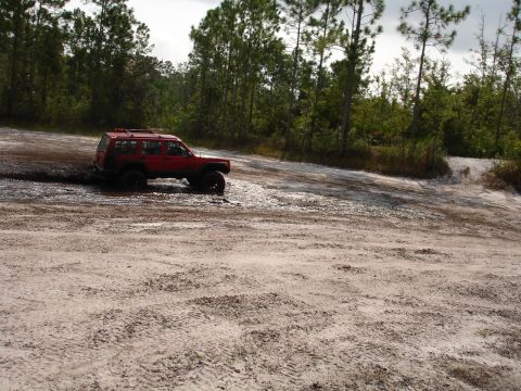 1989 Ford Bronco - A red SUV is stuck in the mud on a dirt path surrounded by trees.