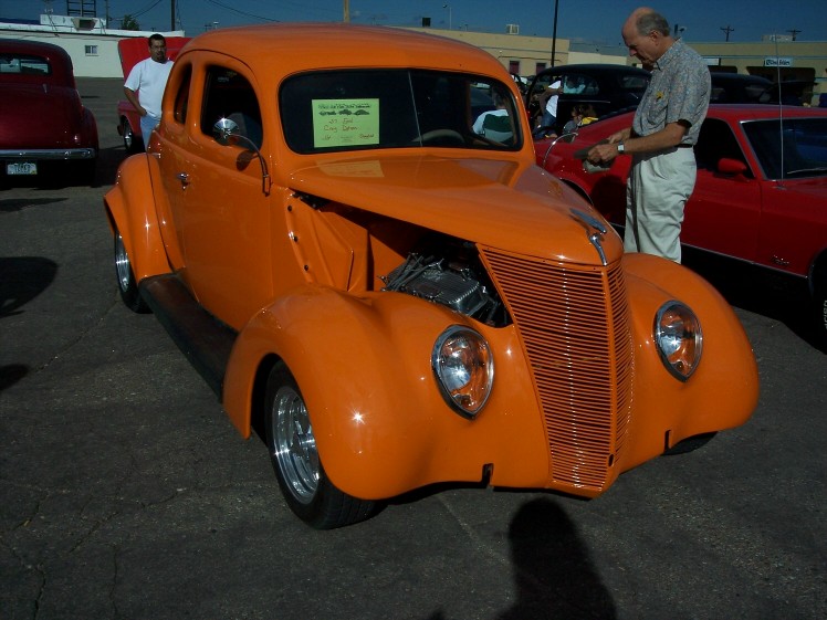 Orange hot rod coupe with hood open, chrome grille, and custom wheels. A sign is on the windshield