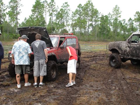 1989 Ford Bronco - Several people inspect a red Jeep Cherokee with its hood up, stuck in a muddy field alongside another heavily soiled truck.