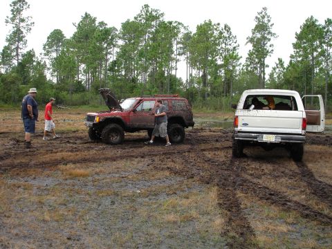 1989 Ford Bronco - A white 1989 Ford Bronco is stuck in the mud with its door open, while another red SUV is being worked on nearby.
