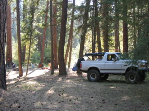 1995 Ford Bronco - A white SUV is parked in a forest clearing, with tents and camping gear visible in the background.