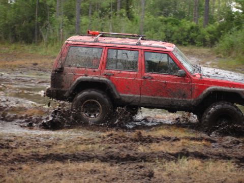 1989 Ford Bronco - This red Jeep Cherokee XJ is navigating a muddy off-road trail, kicking up water.