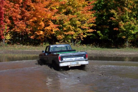 1979 Ford Bronco - A Ford pickup truck navigates a muddy terrain with colorful fall foliage behind it.