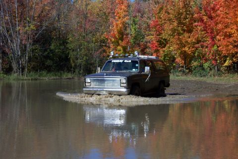 1979 Ford Bronco - A dark SUV navigates through a muddy puddle surrounded by colorful autumn foliage.