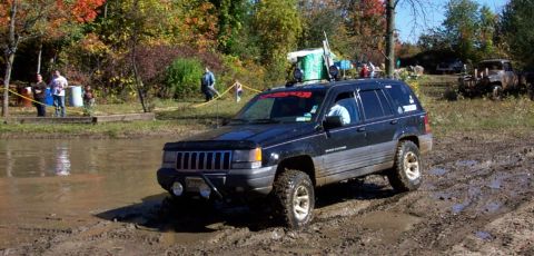 1979 Ford Bronco - A black Jeep Grand Cherokee navigates a muddy off-road course with spectators nearby.