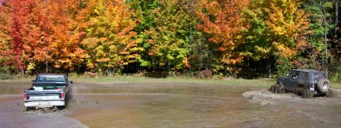 1979 Ford Bronco - Two off-road vehicles navigate a muddy terrain amidst vibrant fall foliage.