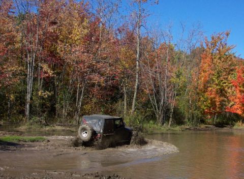 1979 Ford Bronco - A black Jeep Wrangler navigates a muddy trail during autumn with colorful foliage in the background.
