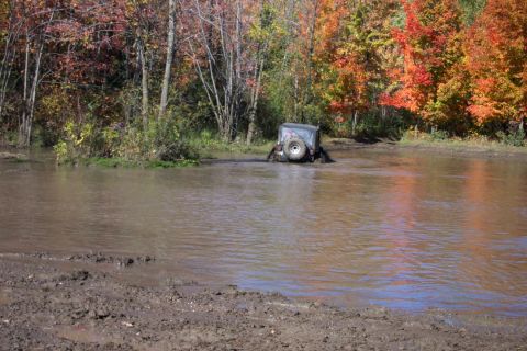 1979 Ford Bronco - An off-road vehicle is partially submerged in muddy water, surrounded by trees with colorful fall leaves.
