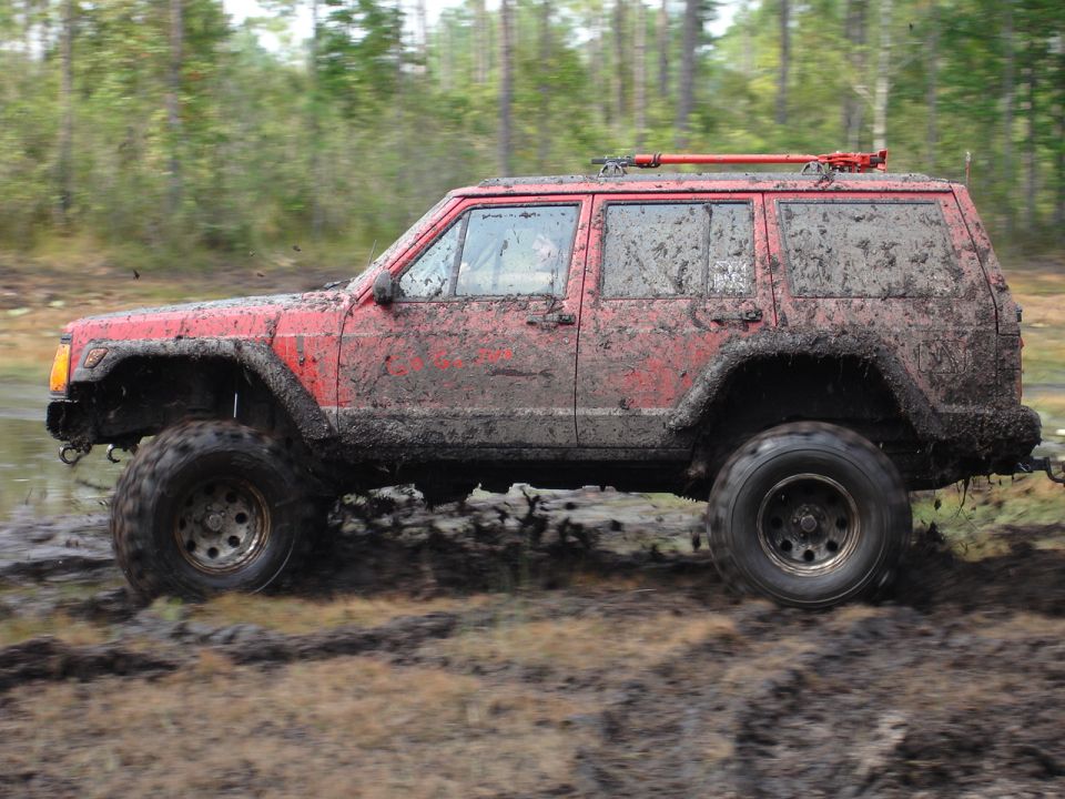 Mud-covered red Jeep Cherokee XJ driving through a muddy trail with trees in the background