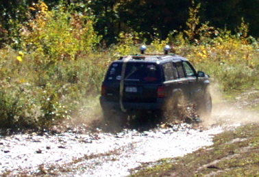 1979 Ford Bronco - A Jeep Grand Cherokee navigates a muddy off-road trail, kicking up dirt and water.