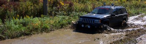 1979 Ford Bronco - A black Jeep Grand Cherokee navigates a muddy trail, kicking up water and dirt.