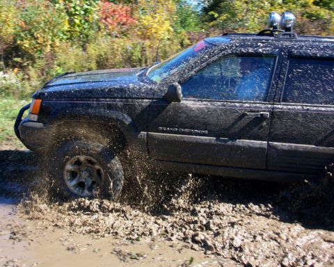 1979 Ford Bronco - A black Jeep Grand Cherokee navigates a muddy trail, kicking up spray as it drives through the mire.
