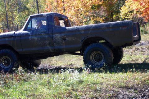 1979 Ford Bronco - A dark, muddy pickup truck navigates a grassy, wooded terrain with fall foliage in the background.