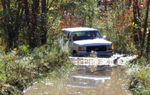 1979 Ford Bronco - A white van navigates through a muddy off-road trail surrounded by trees.