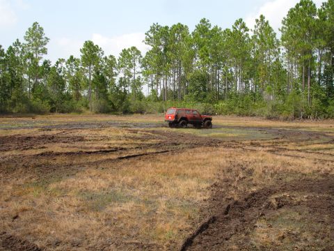 1989 Ford Bronco - A red SUV navigates a muddy terrain with a line of trees in the background.