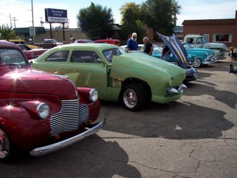 1972 Ford Ranchero - A line of custom cars are on display at a car show, including a green coupe with its hood up.