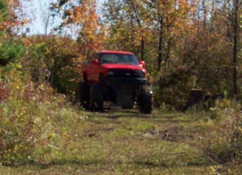 1979 Ford Bronco - A red pickup truck modified with oversized tires drives off-road through a wooded area.