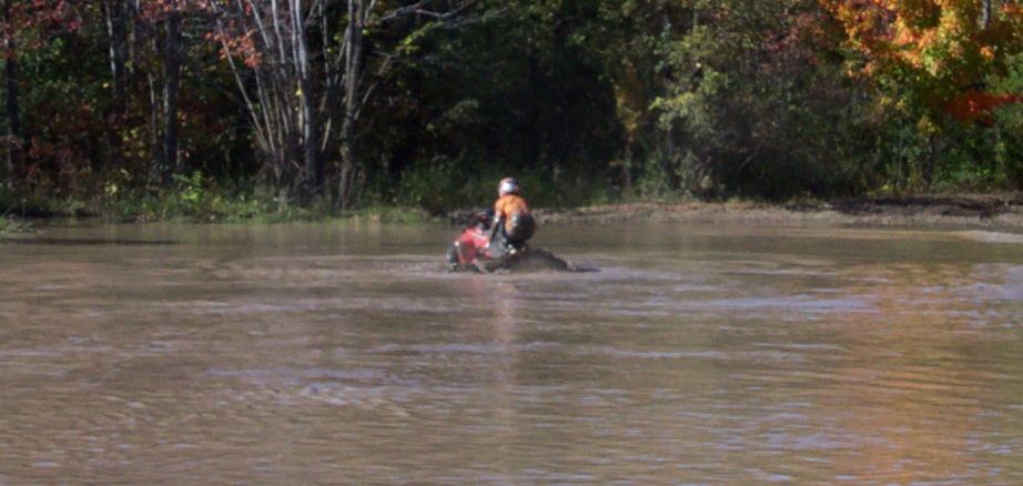 Person on an ATV rides through murky water with trees in the background