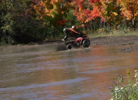 1979 Ford Bronco - A rider navigates a red ATV through a muddy water obstacle with colorful autumn foliage behind.