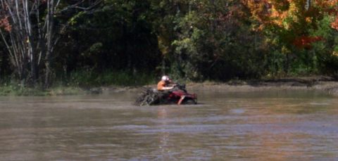 1979 Ford Bronco - A red ATV is stuck in muddy water, with trees and foliage visible on the bank behind it.