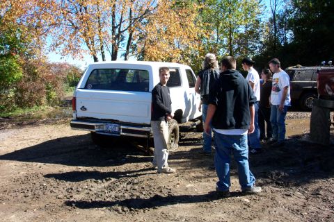 1979 Ford Bronco - A group of people gather around a white SUV parked in a muddy area with autumn trees in the background.