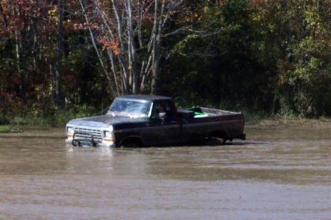 1979 Ford Bronco - A dark-colored pickup truck is seen from the side, driving on a paved surface with trees in the background.
