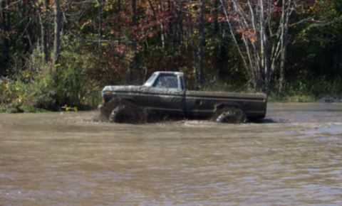 1979 Ford Bronco - A pickup truck is driving through a muddy water crossing with trees in the background.