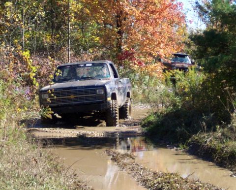 1979 Ford Bronco - Off-road trucks navigate a muddy path surrounded by trees with autumn leaves.