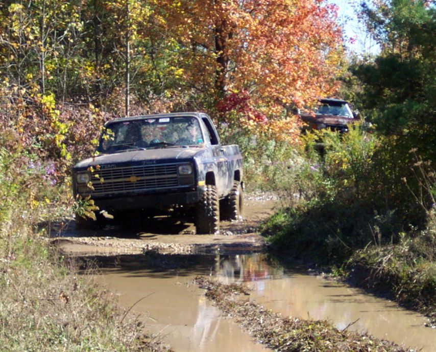Two pickup trucks driving through a muddy trail in the woods with fall foliage