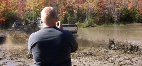 1979 Ford Bronco - Two trucks navigate a muddy obstacle course during autumn, sending plumes of water and mud into the air.