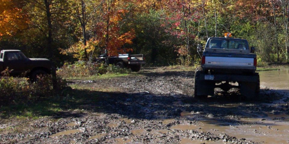 Three trucks driving through a muddy, wooded area during autumn