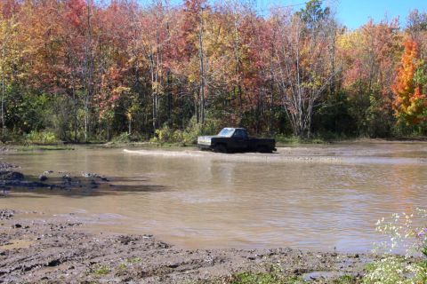 1979 Ford Bronco - Pickup truck navigates a water obstacle during autumn. Supermotors.net logo present.
