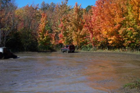 1979 Ford Bronco - Two SUVs are seen in a muddy area surrounded by trees displaying vibrant fall colors.