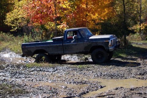 1979 Ford Bronco - A dark pickup truck navigates a muddy terrain with fall foliage overhead.