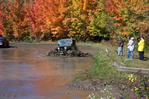 1979 Ford Bronco - An off-road vehicle navigates a muddy trail during autumn, with spectators observing from the bank.