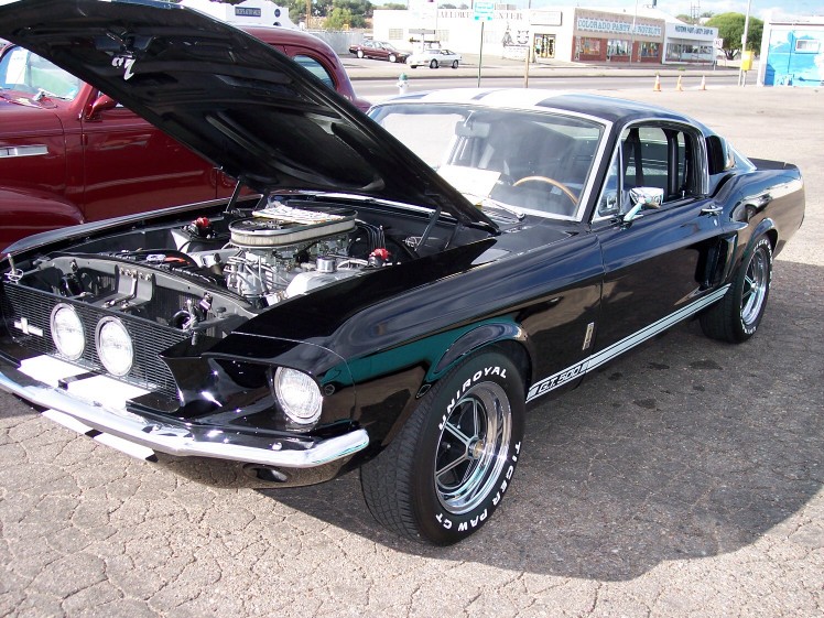 Black 1967 Ford Mustang Shelby GT500 with hood open, showing engine. White racing stripes on hood and roof. Another car is partially visible to the left
