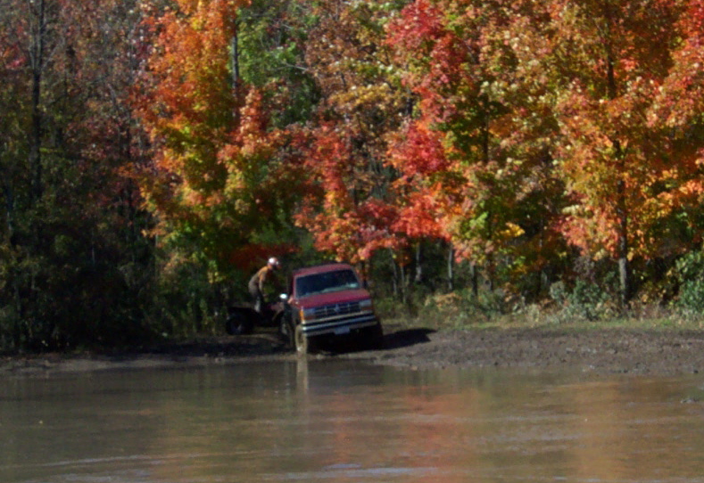 Red pickup truck partially submerged in mud, person on ATV nearby, autumn trees in background